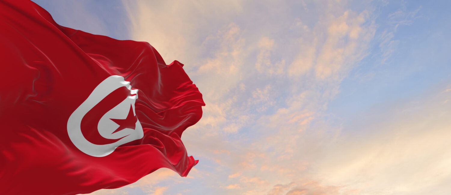 Large flag of Tunisia waving in the wind on flagpole against the sky with clouds on sunny day
